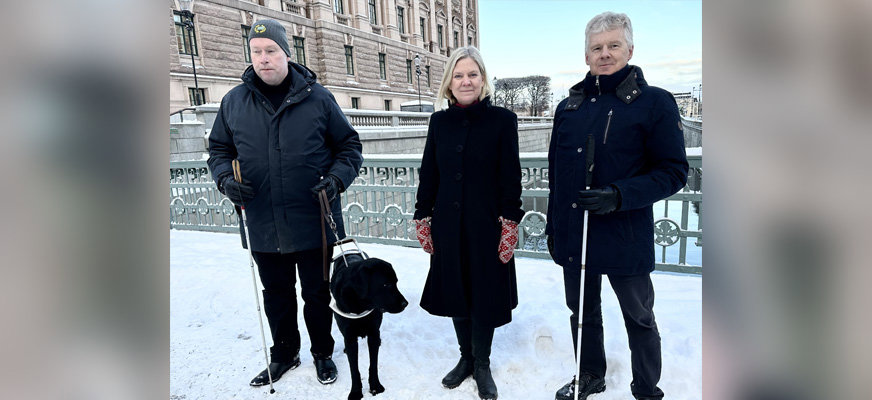 Magdalena Andersson står mellan SRFs ordförande Niklas Mattsson och sakkunnig Jimmy Pettersson, med ledarhunden Quick, på bron utanför riksdagen. Det är snö på marken.