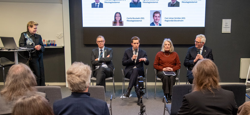 Politikerpanelen bestående av Tomas Ragnarsson (M), Carl-Johan Schiller (KD), Agneta Nilsson (S) och Anders W Johnsson (C) sitter uppradade framför en storbildsskärm. Carl-Johan och Anders håller i mikrofoner och Carj-Johan pratar till åhörarna som sitter framför dem. Bredvid panelen står moderatorn, lutad mot ett bord med en laptop på.