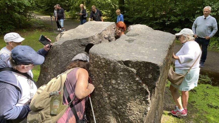 Bilden visar en grupp människor runt ett konstverk i form av en delad sten i ett skogsområde. En person tittar genom en öppning i stenen medan andra undersöker den. Installationen smälter samman konst och natur, vilket väcker intresse och diskussion bland besökarna. Konstverket verkar skapa en interaktiv och utbildande upplevelse.