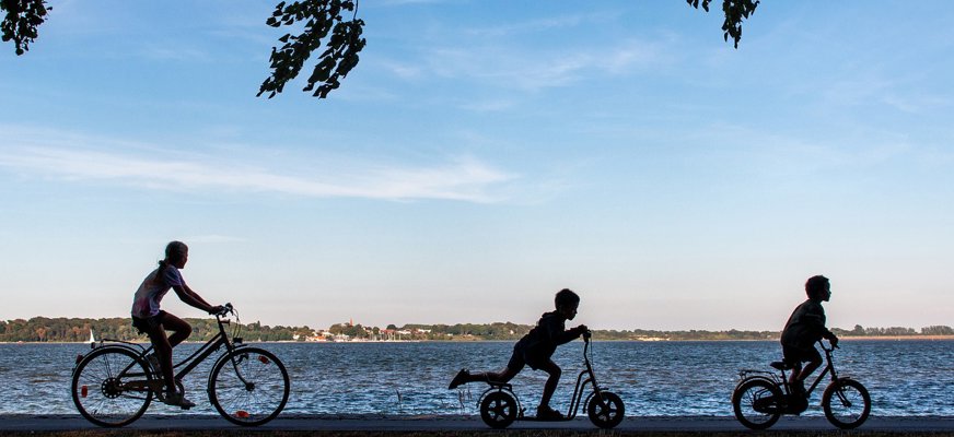 Tre barn cyklar på rad bredvid vatten.