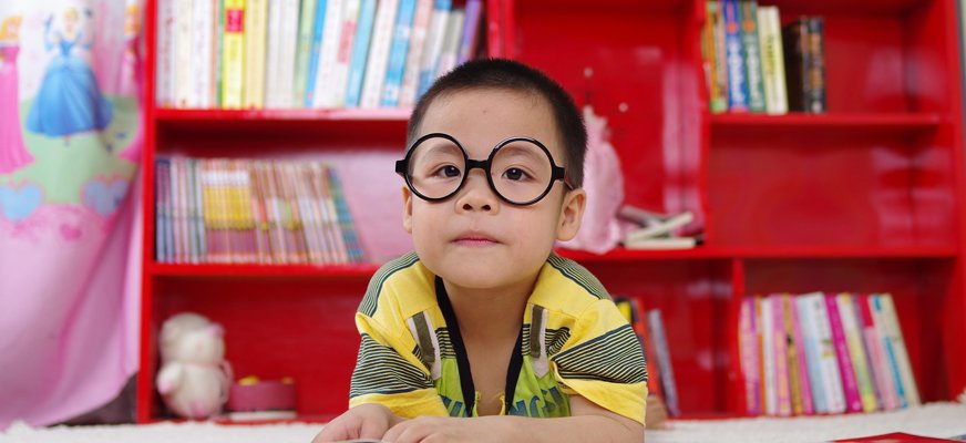 En liten kille svart hår och glasögon ligger på mage och läser en bok. I bakgrunden syns en bokhylla full med böcker.
