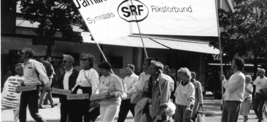 Svartvitt foto demonstrationståg på gata. Stor banderoll med text "Jämlikhet + Rättvisa" Synskadades Riksförbund SRF.