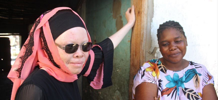 Två personer. Afrikansk kvinna med albinism i sjal, solglasögon, lutar sig i dörröppning. Svart kvinna i blommig tröja bredvid.