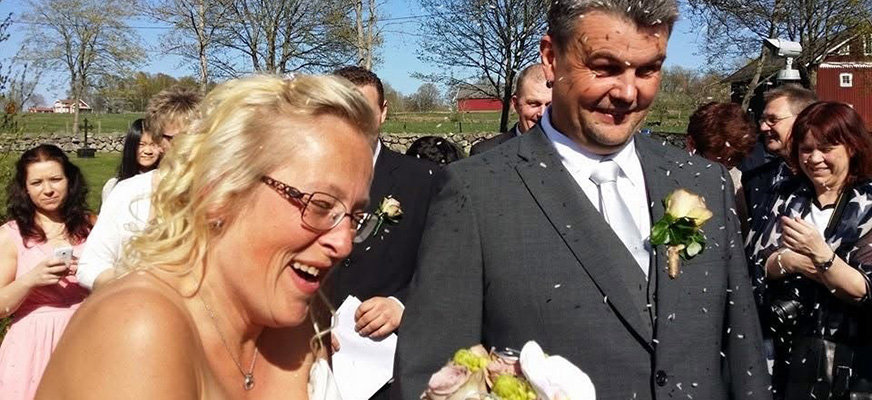 Ett nygift par går utomhus medan ris kastas över dem. De skrattar, och bruden håller en bukett. Flera gäster syns i bakgrunden under klarblå himmel.