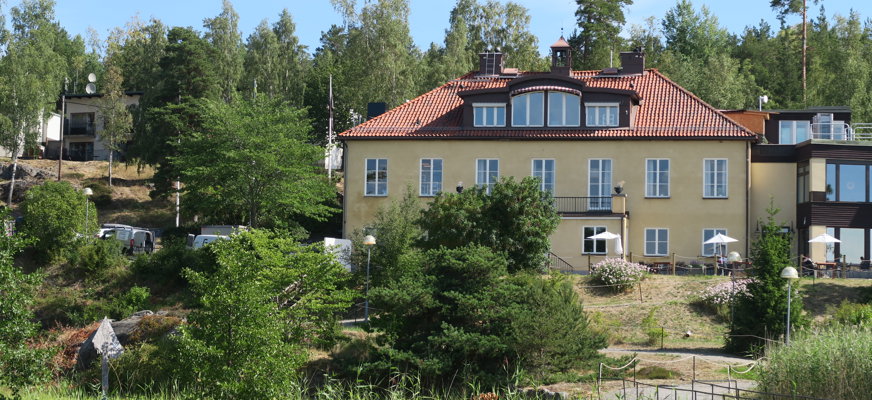 En gul herrgårdsliknande byggnad tronar upp bland grönskande träd och trädgård.