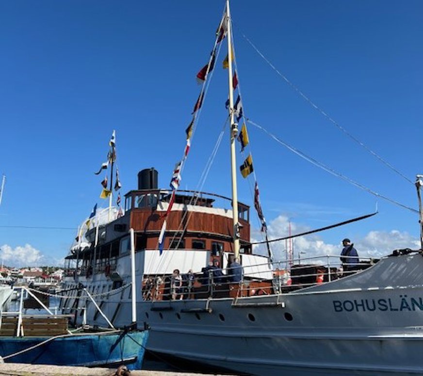 Bilden visar ångbåten med fören mot kajen och på det vita skrovet står namnet "Bohuslän". Uppe på däck syns några besättningsmän klädda i mörkblå kläder. Båten har två master med signalflaggor, en svart skorsten och en rödbrun överbyggnad med fönster.