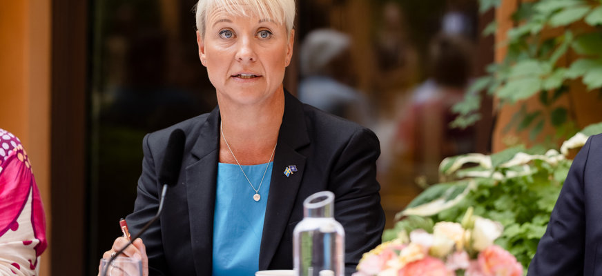 Anna Tenje har kort, blont hår och bär en svart kavaj med en svenska flaggan-brosch på kragen. Hon sitter vid ett bord och i förgrunden syns en vas med blommor och en karaff med vatten.