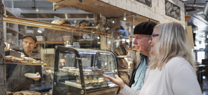 Kvinna syntolkar konditoridisk för man med vit käpp.