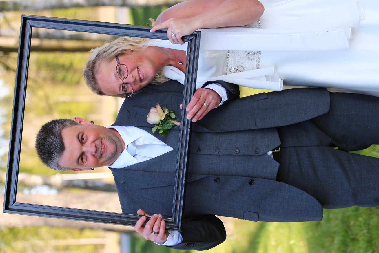 Ett brudpar står utomhus och ler mot kameran. De håller upp en tom tavelram framför sig. Personen till vänster bär grå kostym med vit slips och blomma på slaget, och personen till höger bär vit bröllopsklänning.