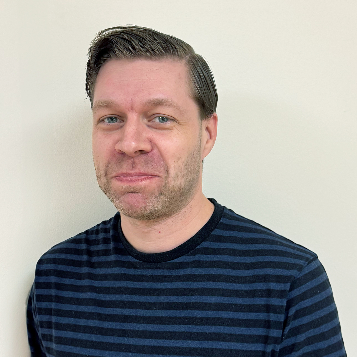 En leende man med brunt hår, blå ögon och skäggstubb, iförd blå- och svartrandig tröja.