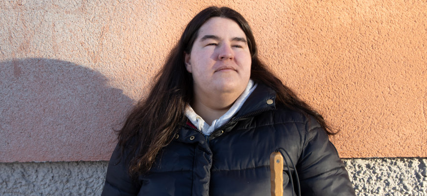 Porträttbild på Oliwia Wisniewska, en ung kvinna i mörkt hår som faller över axlarna. Oliwia står i solen, mot en byggnad med putsad fasad i orange. Hon har en allvarlig uppsyn och blickar uppåt.