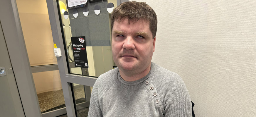 Porträttbild på Damir. Han sitter ute i en korridor på vad som ser ut som en mottagning av något slag. Damir har kort brunt hår, skäggstubb och är klädd i en grå stickad tröja över t-shirt. Det är tydligt på hans ögon att han är synskadad.