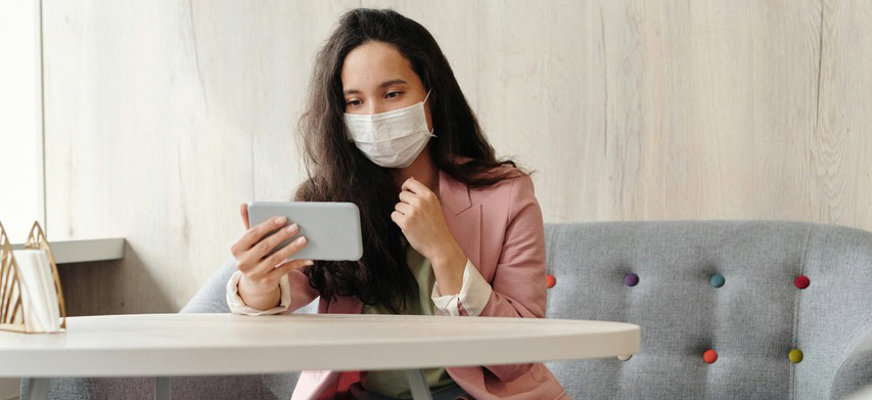 En kvinna med munskydd sitter i en soffa och håller telefonen framför sig, som om hon har ett videosamtal.