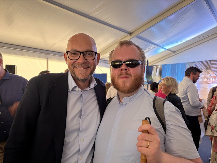 Linus Forsberg från SRF:s förbundsstyrelse poserar tillsammans med arbetsmarknadsminister Johan Britz (L). Johan är flintskallig, har kort skägg och bär glasögon med svarta bågar. Han bär ljusblå skjorta med mörk kavaj över. Linus är tunnhårig med rödlätt skägg och bär en ljusblå skjorta och svarta solglasögon. I handen har han en vit käpp.