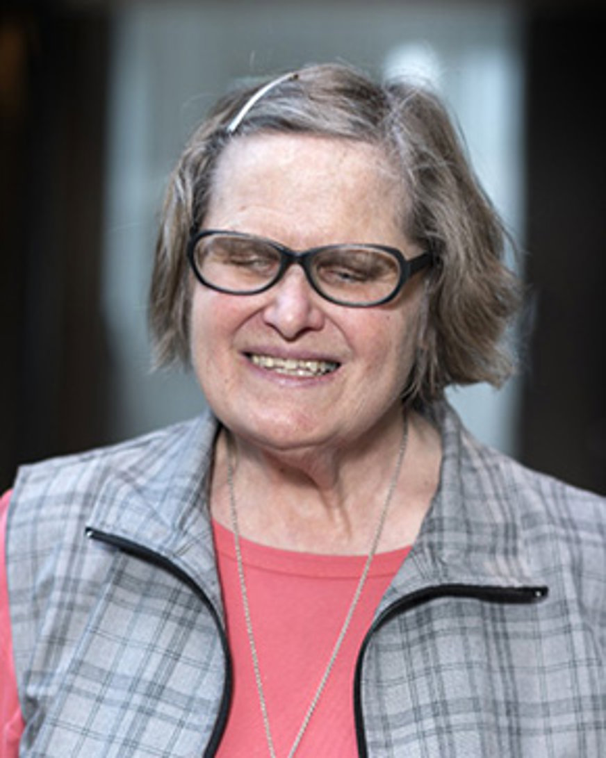 Porträttbild Anita Svenningsson: Kvinna med kort gråspräckligt hår iförd röd tröja med grårutig väst över. Hon har ett hårspänne i håret och bär ovala glasögon med grova, svarta bågar.