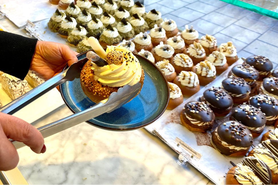På insidan av skyltfönster på ett bageri, ser man flera semlor av olika slag. Händer syns i förgrunden, den ena håller i en talrik den andra i en tång med en semla ovanför tallriken.