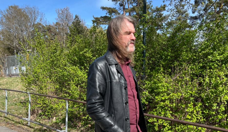 Fredrik, i långt gråbrunt hår och skägg går utomhus i en svart skinnjacka över en vinröd skjorta. Han går framför grönskande buskar och träd i solsken.