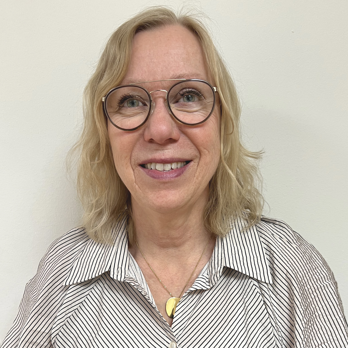 Leende kvinna 60-årsåldern, blont axellångt hår, glasögon, randig skjorta.