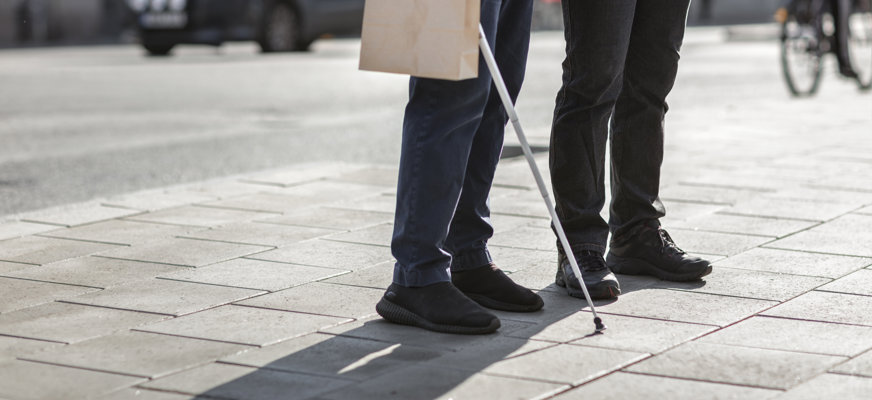 En person med vit käpp blir ledsagad i en gatumiljö.