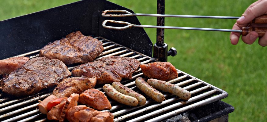 Bilden visar en grill med hamburgare, köttbitar och korvar. Från höger kommer en hand som håller i en grilltång in i bilden.