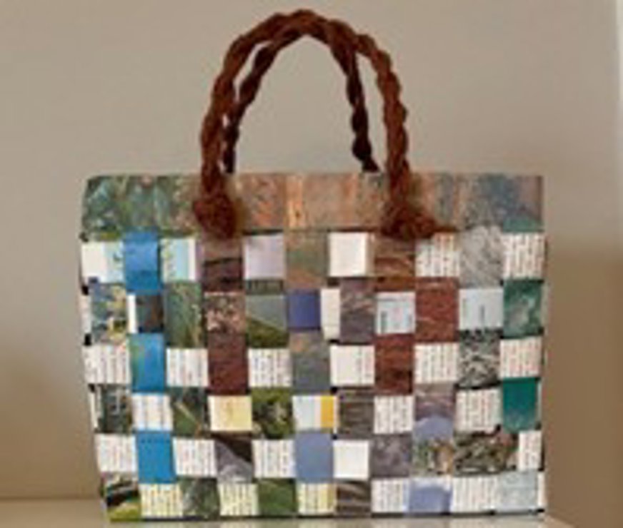 Väskan är rektangulär och handgjord av flätade remsor från tidningar eller magasin. Remsorna bildar ett rutmönster i varierande färger som blått, grönt, brunt och vitt, med synlig text på flera av dem. Handtagen är två tjocka, tvinnade snören i brun färg, fästa upptill på väskan. Den har ett rustikt och kreativt uttryck, där återbruk av material ger både funktion och dekorativt värde. Bakgrunden är neutral och framhäver väskans detaljer.