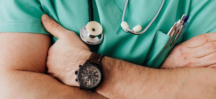 Närbild på en man som korsar sina armar framför bröstet. Han har på sig läkarrock och ett stetoskop hänger runt halsen.