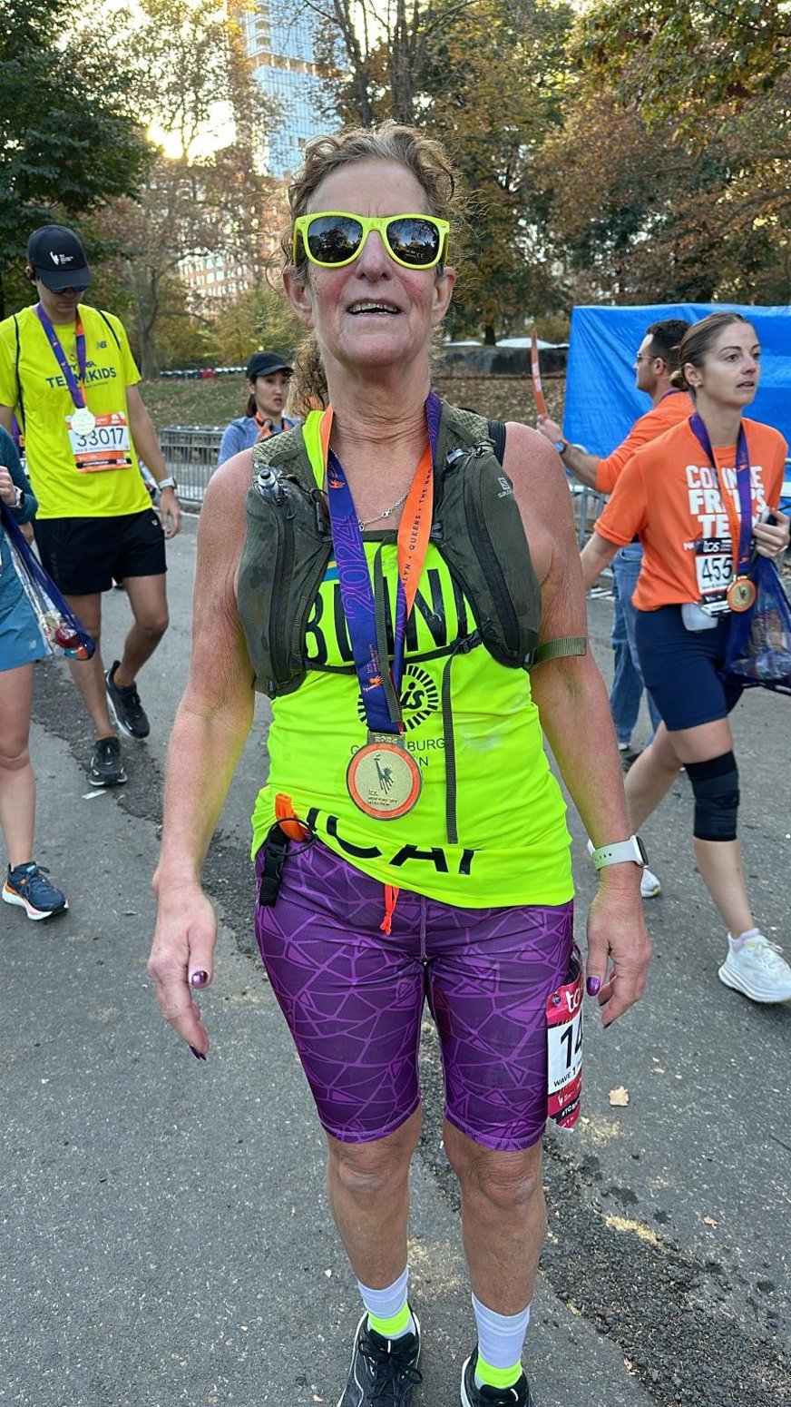 Denise ser lycklig och lättad ut med en medalj runt halsen. Hon har solglasögon med gula bågar, ett gult linne med texten BLIND, lila korta tajts och en löparryggsäck. Bakom henne skymtar andra löpare med medalj runt halsen.