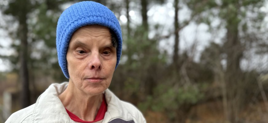 Porträttbild på Eva Nilsson som har blå mössa och vit jacka. I bakgrunden höstlandskap med träd och brunt gräs.