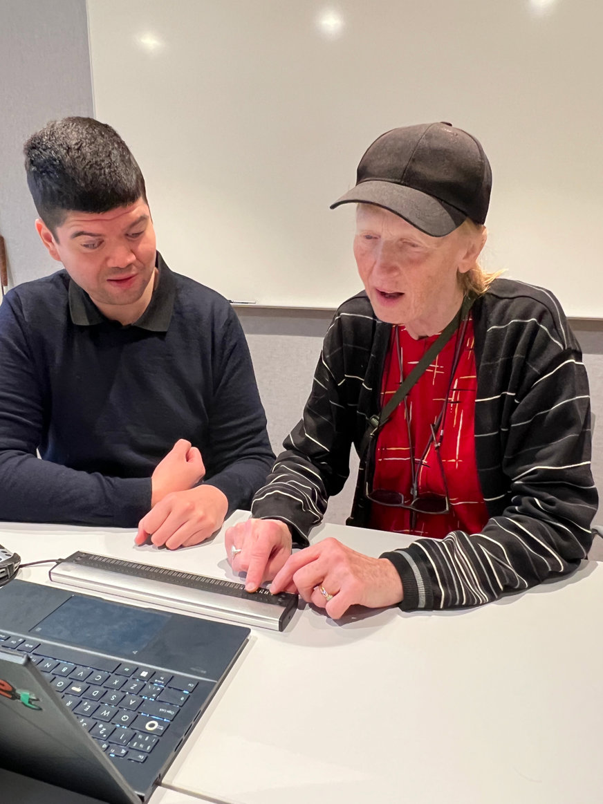 Claudio instruerar Agneta Hummelhielm att använda punktdisplay till en dator. Claudio är kortklippt med mörkt hår och är klädd i svarta kläder. Agneta har en röd tröja under en svart kofta och en svart keps med skärm.. 