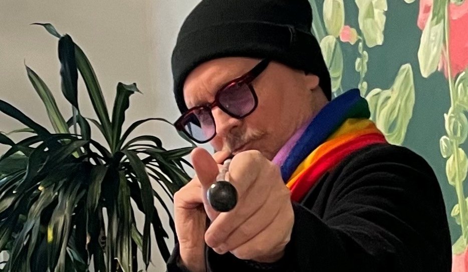 Mathias pekar mot kameran med sin vita käpp, som om han siktar med en biljardkö. Han är en medelålders man med mustasch som bär svarta glasögon med kraftiga bågar och tonade glas. Han har på sig en svart mössa och tröja, och bär en regnbågsrandig halsduk.