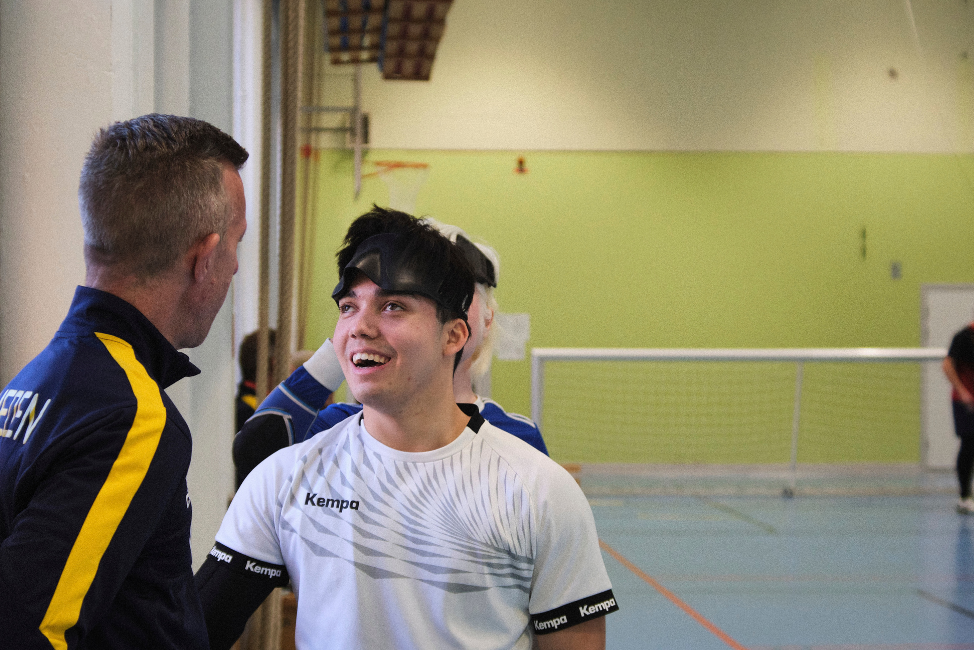 En ung vit man i en vit träningströja pratar med en medelålders vit man i träningsoverallsjacka. Den unge mannen ler och har en svart ögonmask uppdragen i pannan. Platsen är en gymnastikhall och i bakgrunden syns ett goalball-mål