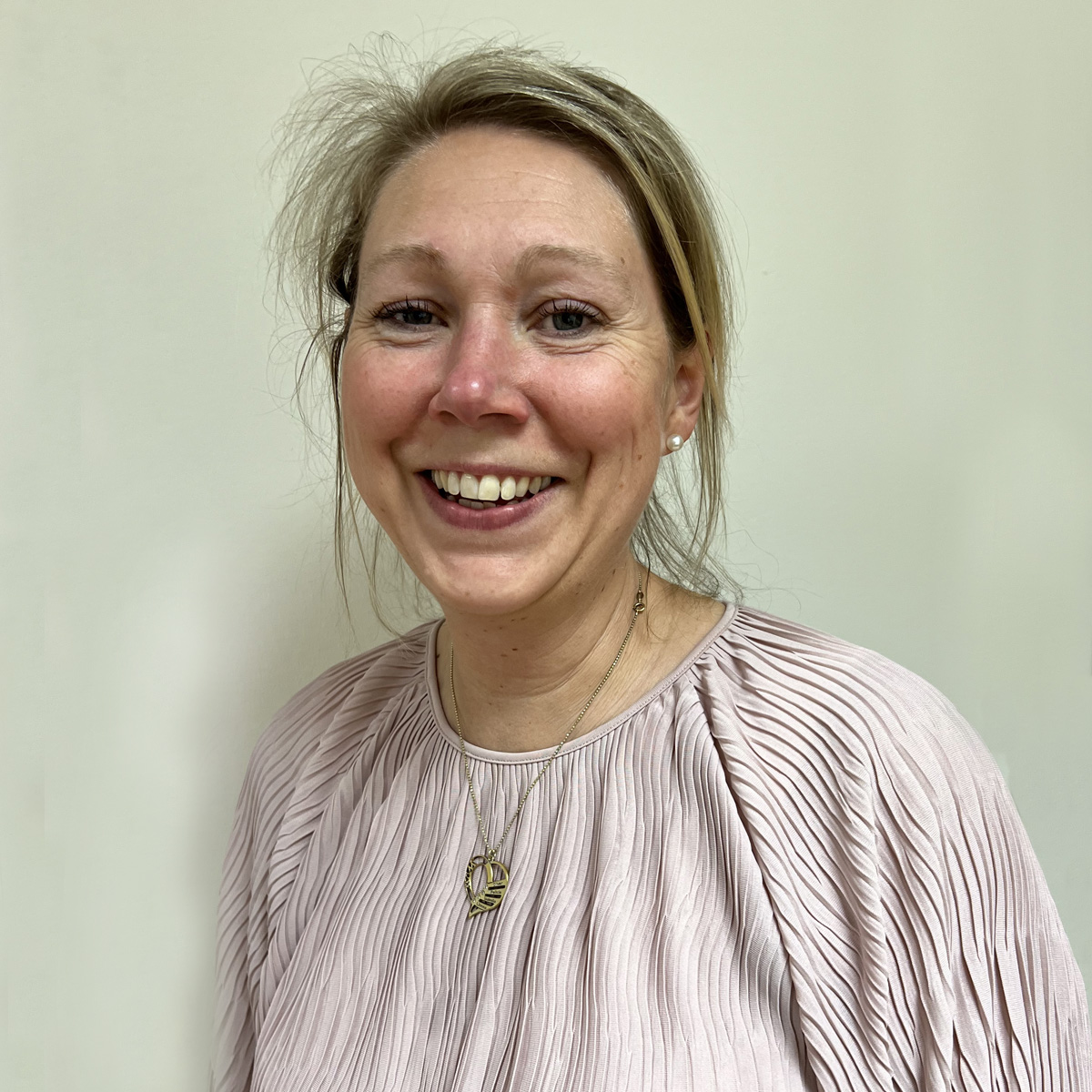 Kvinna i 40-årsåldern, stort leende, blont uppsatt hår, skrattrynkor, plisserad ljusrosa blus, guldhalsband.