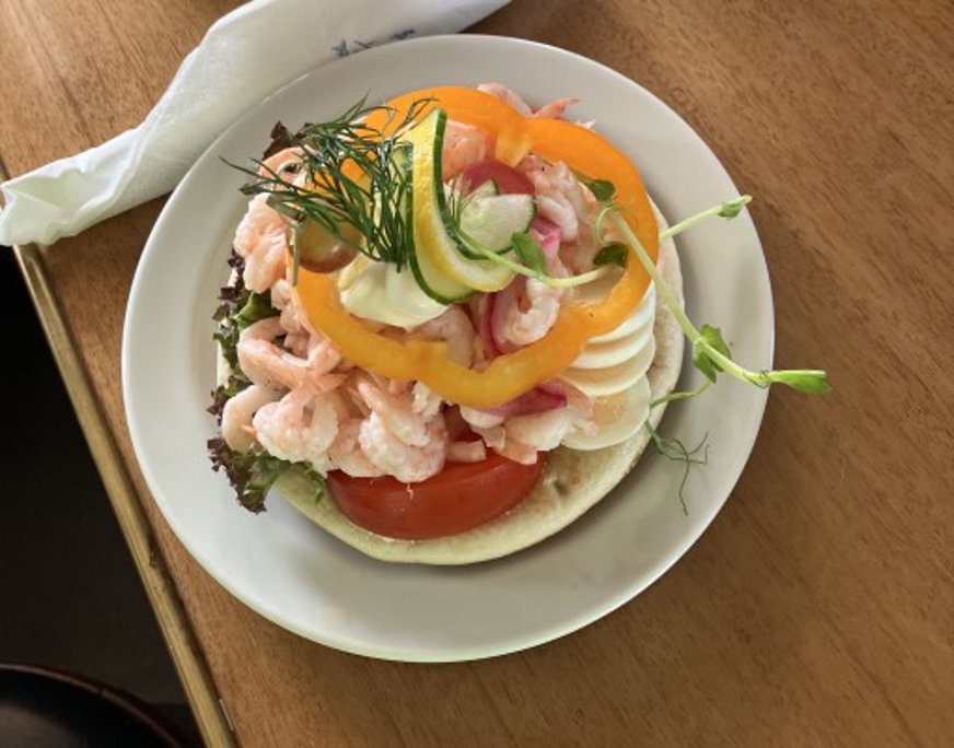 En räksmörgås på en vit tallrik som står på ett träbord. Smörgåsen är toppad med räkor, skivade ägg, tomat, gul paprika, rödlök, en dillkvist och en bit gurka.