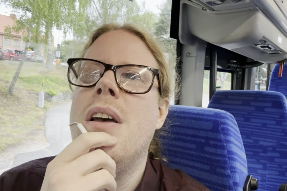 En vit man i 40-årsåldern sitter på en buss med blå säten, han har glasögon och håller en vit sladd med mikrofon vid munnen. Han har blont långt hår uppsatt i en hästsvans. Bussens tomma säten och träd syns i bakgrunden.