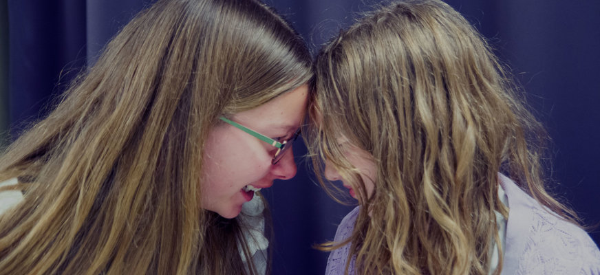 Två unga tonårstjejer i profil, lutar pannorna mot varandra och ler.