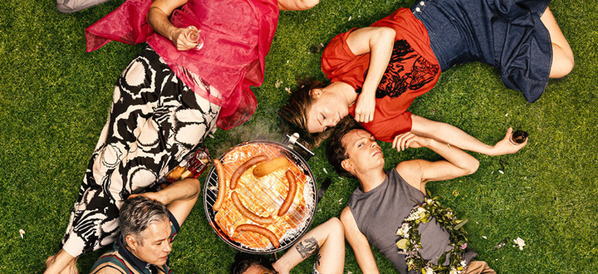 Flera personer ligger på en gräsmatta runt omkring en grill med korvar på. Någon äter på en korv, en annan dricker ur ett champagneglas