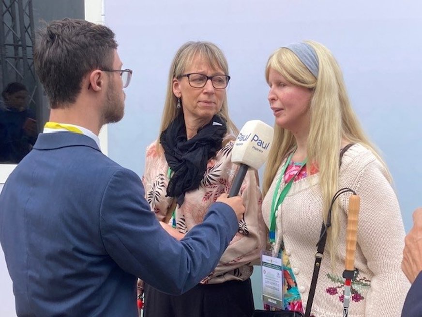 Blond kvinna med långt hår blir intervjuad av man i kostym som håller en mikrofon mot henne. I bakgrunden medelålders kvinna som tittar på.