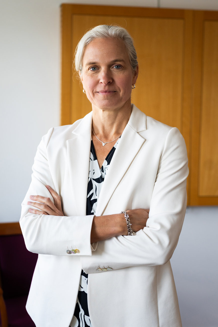 Jenny står inomhus med armarna i kors och ler mjukt mot kameran. Hon har vit kavaj med en svartvitmönstrad blus under. Håret är vitt och uppsatt i nacken och hennes ögonbryn är mörka. Jenny har armband och halsband i silver och matchande ringar i öronen.