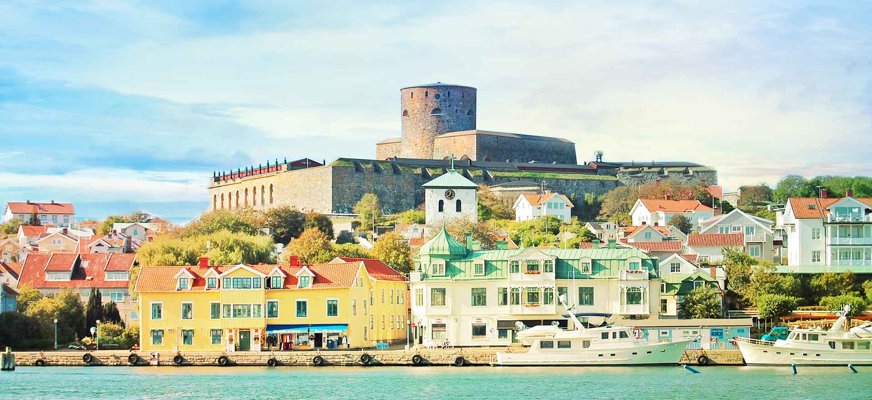 Utsikt över hus vid vattnet i Marstrand, med båtar förtöjda i förgrunden och Carlstens fästning som reser sig ståtligt på höjden i bakgrunden under en ljusblå himmel.