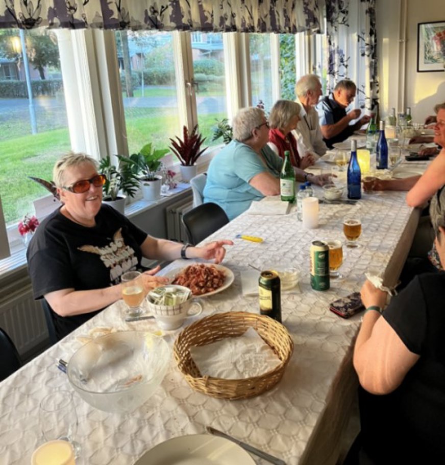 Bilden visar människor som sitter runt ett långbord inomhus. Det är en ljus och solig dag. På bordet ligger en vit duk och det finns tallrikar med mat och olika drycker i flaskor och glas. En kvinna i förgrunden ler mot kameran. Hon har en tallrik med räkor framför sig. I bakgrunden syns stora fönster med blommiga gardiner och utanför fönstren kan man se en grön gräsmatta och några träd.