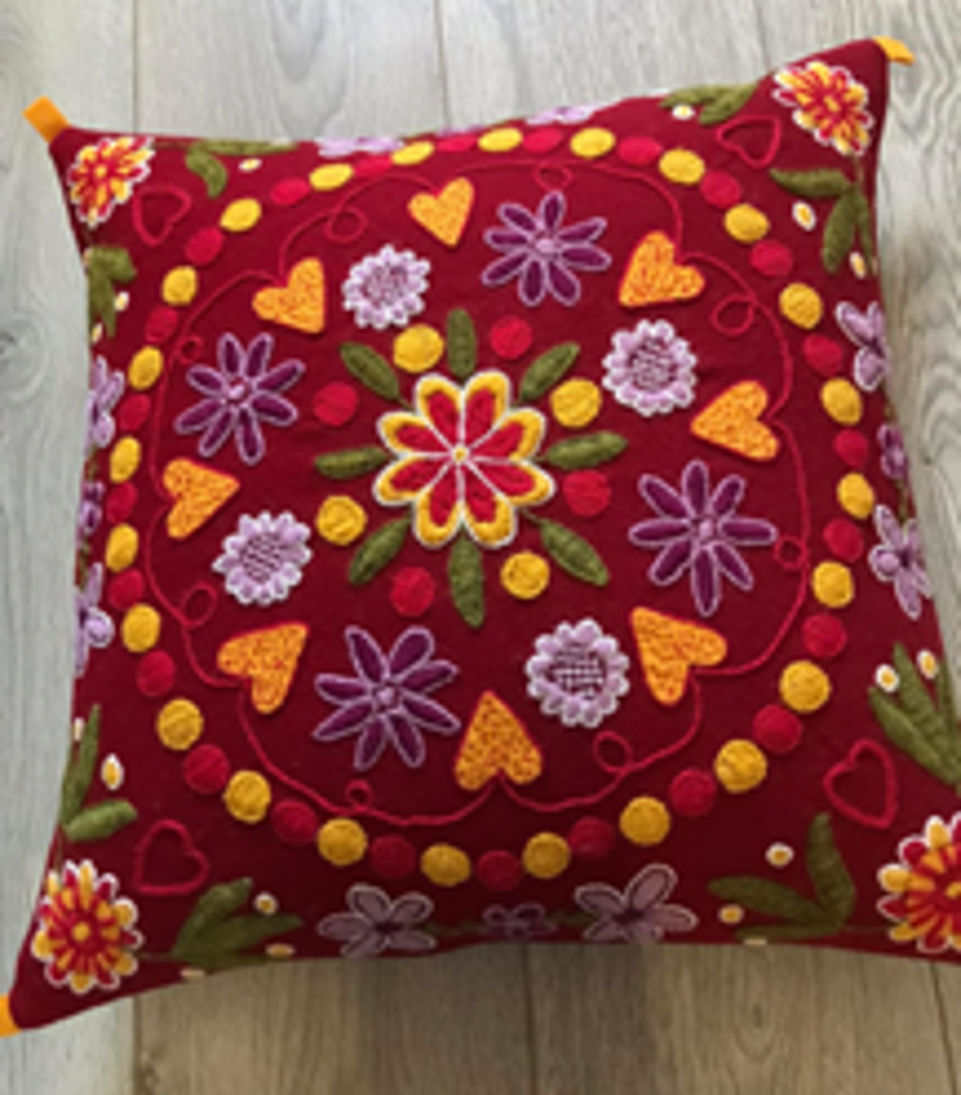 Kudden är fyrkantig med en intensivt röd tygbotten och pryds av riklig brodyr i ett symmetriskt mönster. I centrum finns en stor blomma med kronblad i gult, rött och vitt, omgiven av mindre blommor i lila, orange och vitt. Runtom syns gröna blad och färgglada prickar i gult och rött som förstärker det livliga uttrycket. Brodyren strålar ut från mitten och skapar en harmonisk och balanserad helhet. Detaljerna är noggrant utförda och ger ett folkloristiskt eller traditionellt intryck, där färg och form samspelar för att skapa en varm och dekorativ textil.