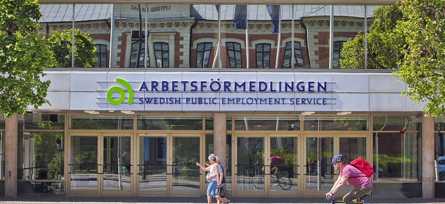 Exteriör bild på ett av Arbetsförmedlingens kontor. Myndighetens logotyp tar stor plats på fasaden. Framför går och cyklar människor.