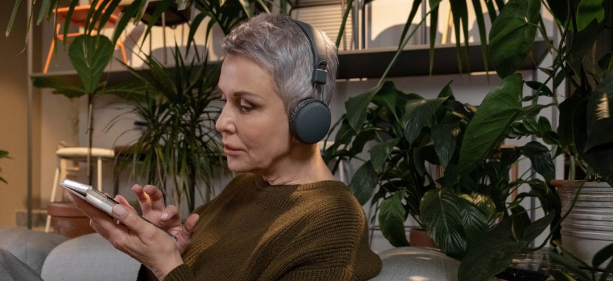 En person med kort grått hår sitter i en fåtölj och använder en mobiltelefon. Personen bär hörlurar och omges av flera stora gröna krukväxter i en hemlik miljö.