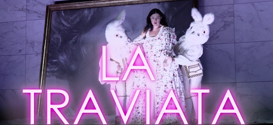 Kvinna i blommig klänning står mellan två personer utklädda till vita kaniner. Framför dem lyser texten ”LA TRAVIATA” i rosa neon.