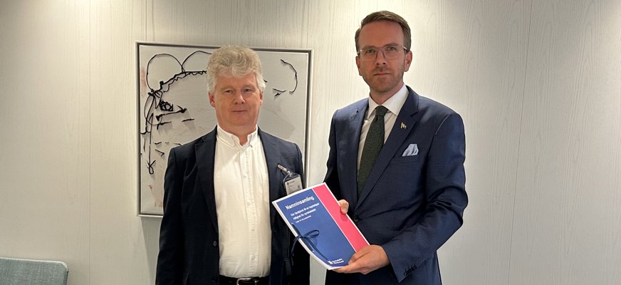 Niklas Mattsson lämnar över namninsamlingen till Andreas Carlson. Båda männen har mörka kavajer och vit skjorta och befinner sig i ett mötesrum på regeringskansliet. Bakom dem skymtar en tavla med ett svartvitt mönster.