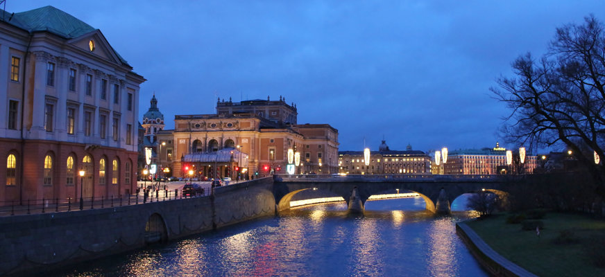 En bild av Stockholms ström mellan Gamla stan och Slussen i skymningsljus, längre bak syns en bro, ett träd med karga grenar och upplysta byggnader.