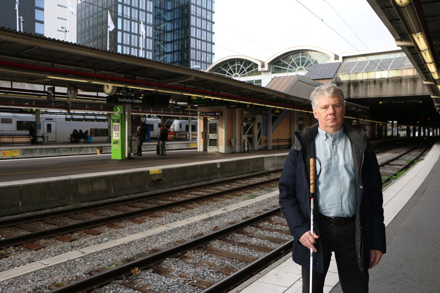 Niklas Mattsson står på en perrong bredvid en tågräls. Han har vit käpp och är klädd i jeans, skjorta och mörkblå jacka. Niklas ser med en allvarlig uppsyn rakt in i kameran.