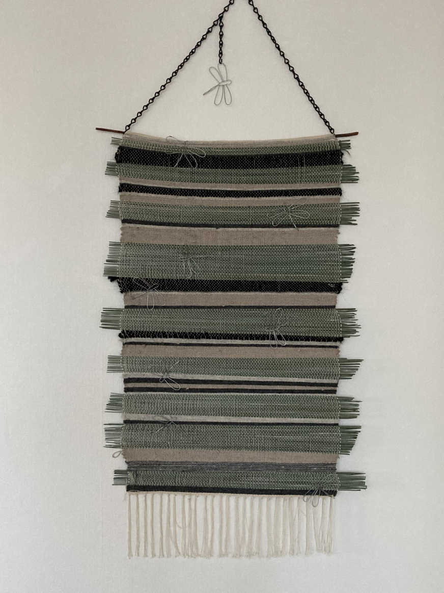 Väggbonaden är ett handvävt textilverk i gröna, grå och vita nyanser, upphängt på en mörk trästav med svart metallkedja. Verket består av horisontella band med varierande texturer, där vissa partier innehåller knutar och dekorativa inslag. Den nedre delen avslutas med en vit frans som ger rörelse och mjukhet. Designen är symmetrisk och välbalanserad, vilket framhäver hantverkets precision och omsorg. Kombinationen av färgskala, struktur och material skapar ett lugnt och stilrent uttryck som passar både som konstverk och inredningsdetalj.