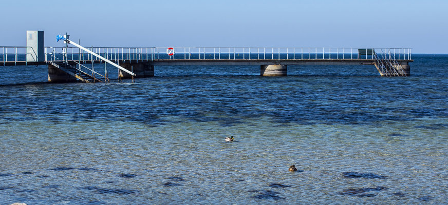 Bilden visar bryggan sedd från stranden, den är vitmålad och det hänger en frälsarkrans på mitten av bryggan.