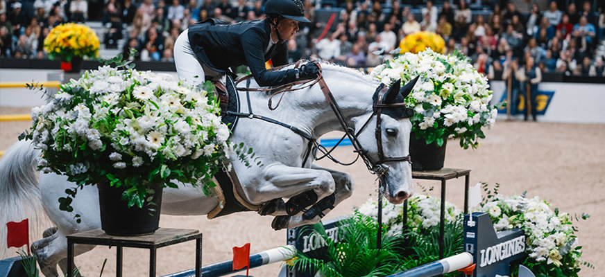 Bilden visar en ryttare klädd i svart hjälm och tävlingsklädsel som rider en vit häst över ett hinder i en hoppningstävling. Hindret är dekorerat med stora blomsterarrangemang i vitt och grönt. Publiken sitter i bakgrunden på läktare och ser på tävlingen. Arenan är inomhus med en ljus sandbana. Ryttaren lutar sig framåt och har en fokuserad hållning medan hästen sträcker ut sig i språnget.